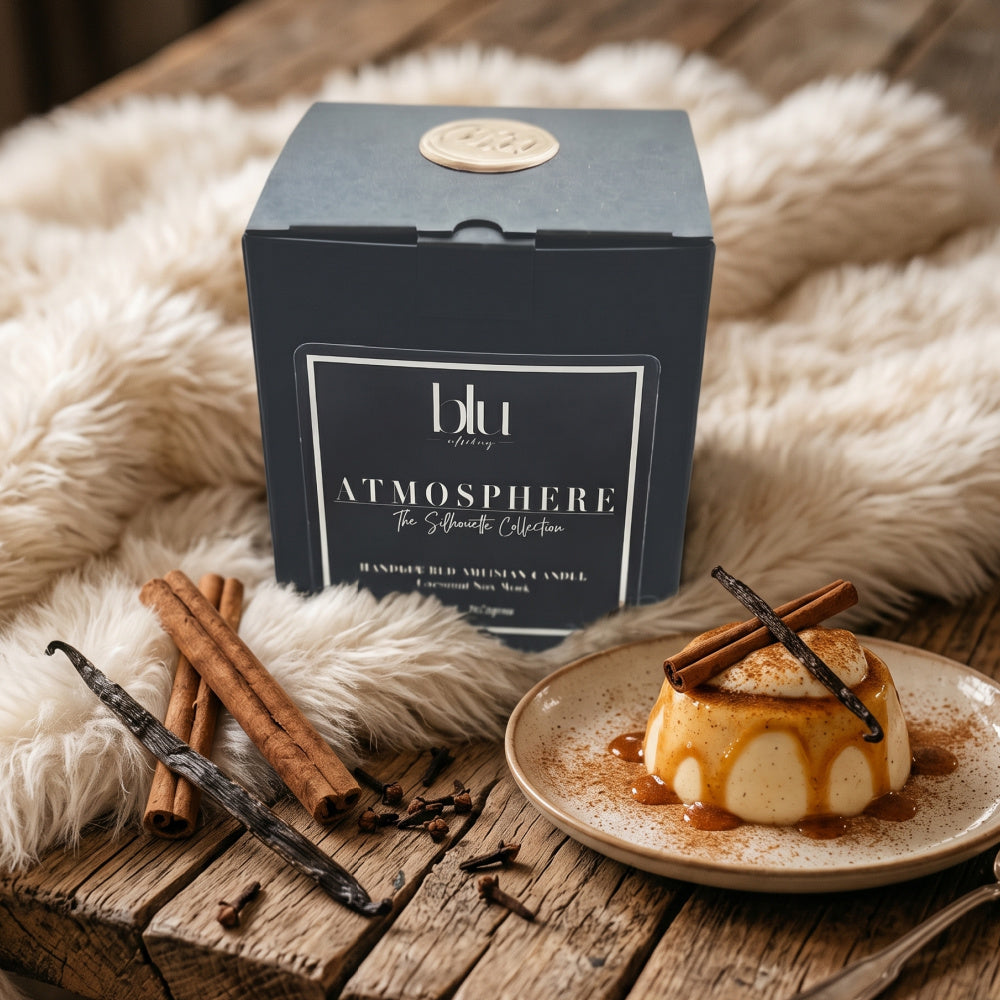The ATMOSPHERE Linger Artisan Candle and its matte black packaging box by BLU, styled on a wooden table with cinnamon, vanilla beans, and a flan dessert in a warm, cozy living room.