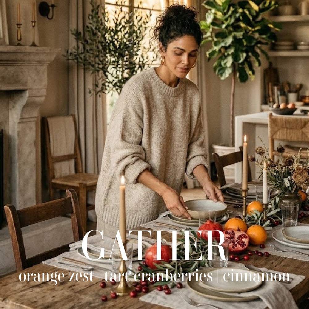 Woman setting a rustic holiday dining table styled with fresh autumn fruit, tapered candles, and the Gather seasonal scent theme