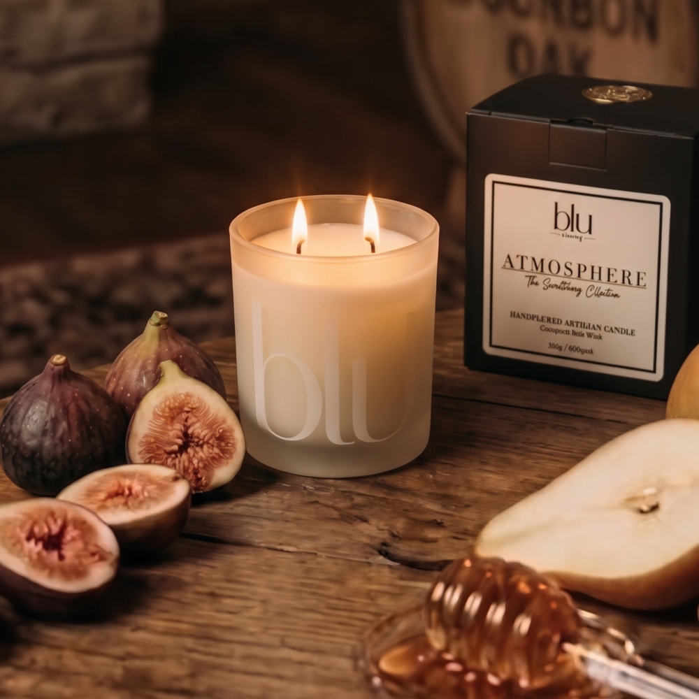 A lit frosted glass candle with two flames sits on a rustic wooden table beside its black branded box, with figs, a pear slice, and honey dipper props in the scene. clean-burning fig bourbon coconut soy candle