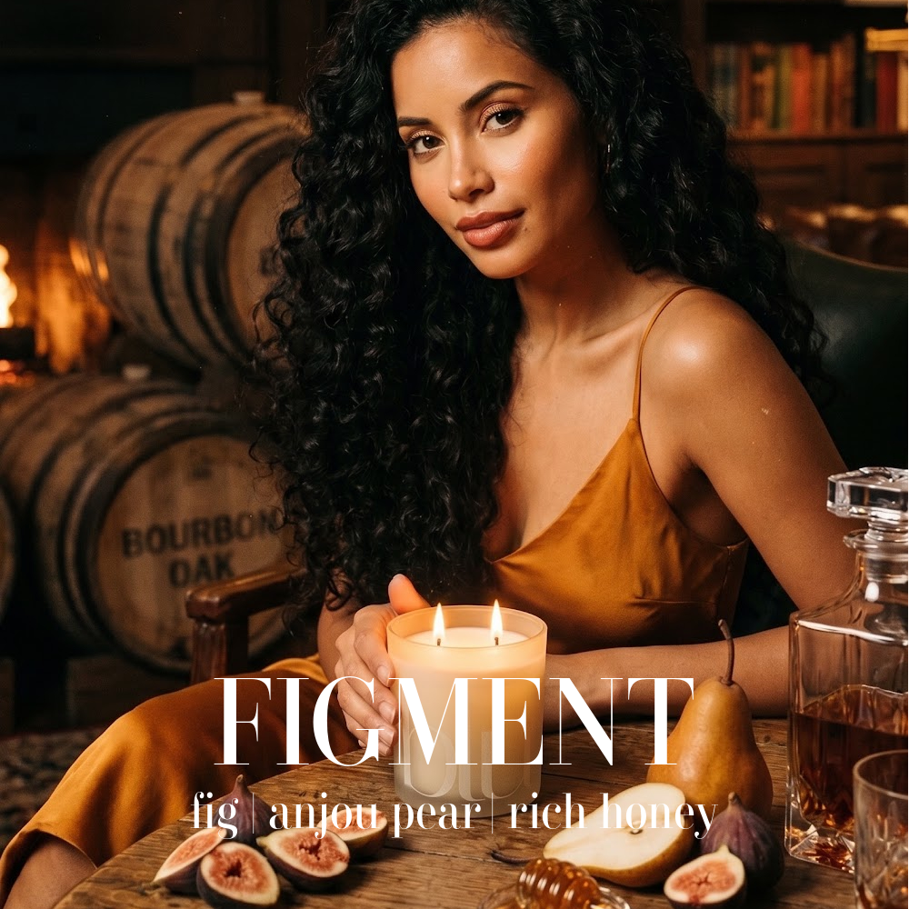 A woman with long curly dark hair sits at a wooden table in a warm, dimly lit room with bourbon barrels in the background. She is wearing a mustard yellow satin camisole and holding a lit candle. figment artisan candle