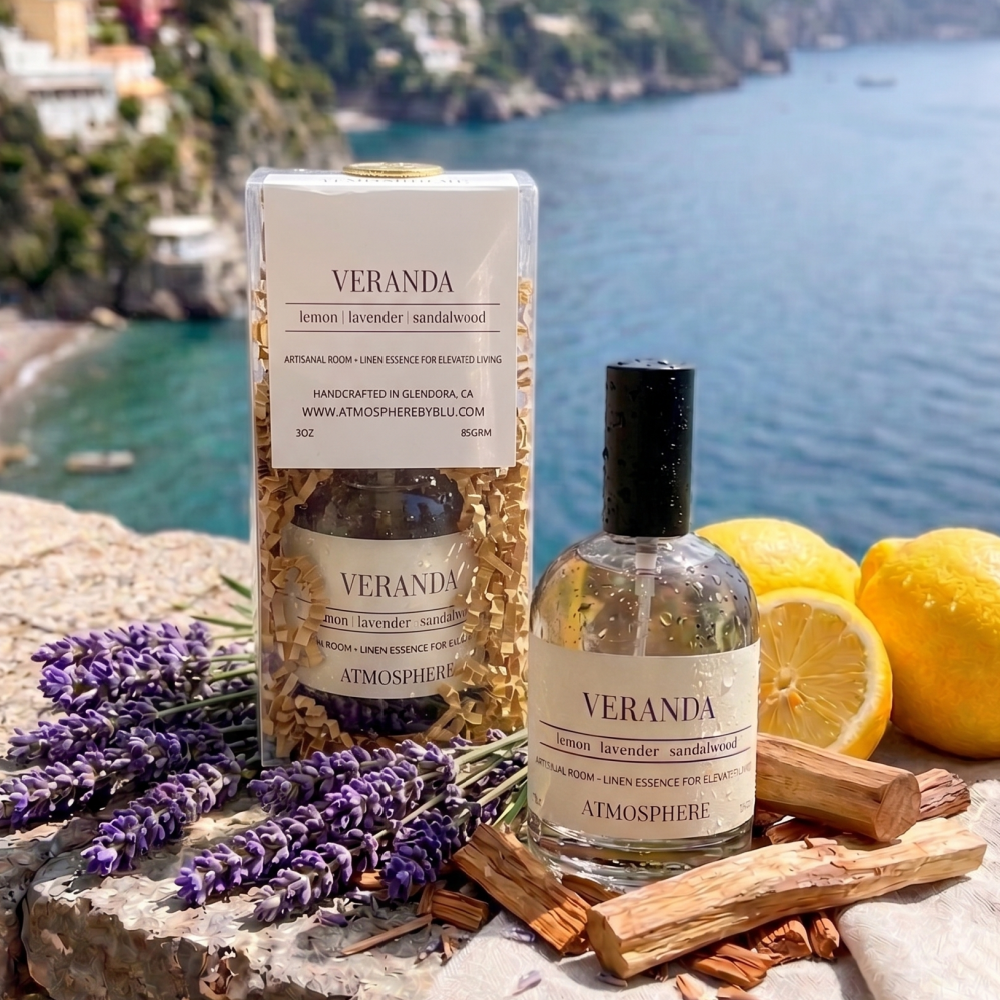 Veranda atmospheric diffuser set featuring a clear cylindrical jar filled with dried botanicals and a matching glass bottle with a black cap, styled on a stone surface with lavender, lemons, and wood pieces against a scenic coastal background. linen mist