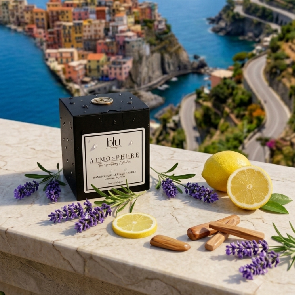 A black square candle tin labeled 'blu Atmosphere' sits on a light stone ledge with lemons, lavender sprigs, and cinnamon sticks, with a blurred coastal town and blue sea in the background. lemon lavender sandalwood candle