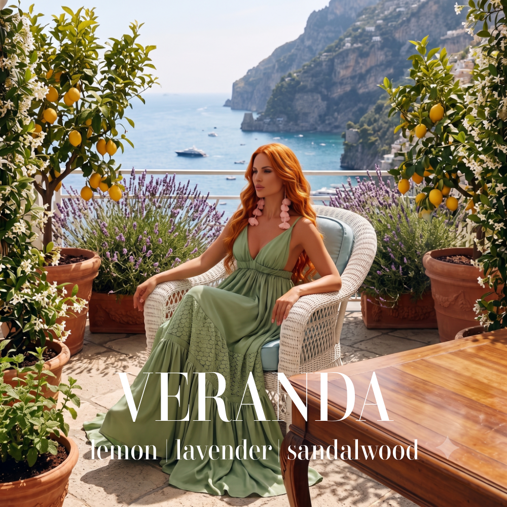 A woman with red hair sits in a white wicker chair on a sunlit terrace overlooking the sea, wearing a flowing sage green dress. Lemon trees and lavender plants frame the scene, with the word 'VERANDA' and the scent notes 'lemon', 'lavender', and 'sandalwood' overlaid at the bottom. Veranda Artisan Candle