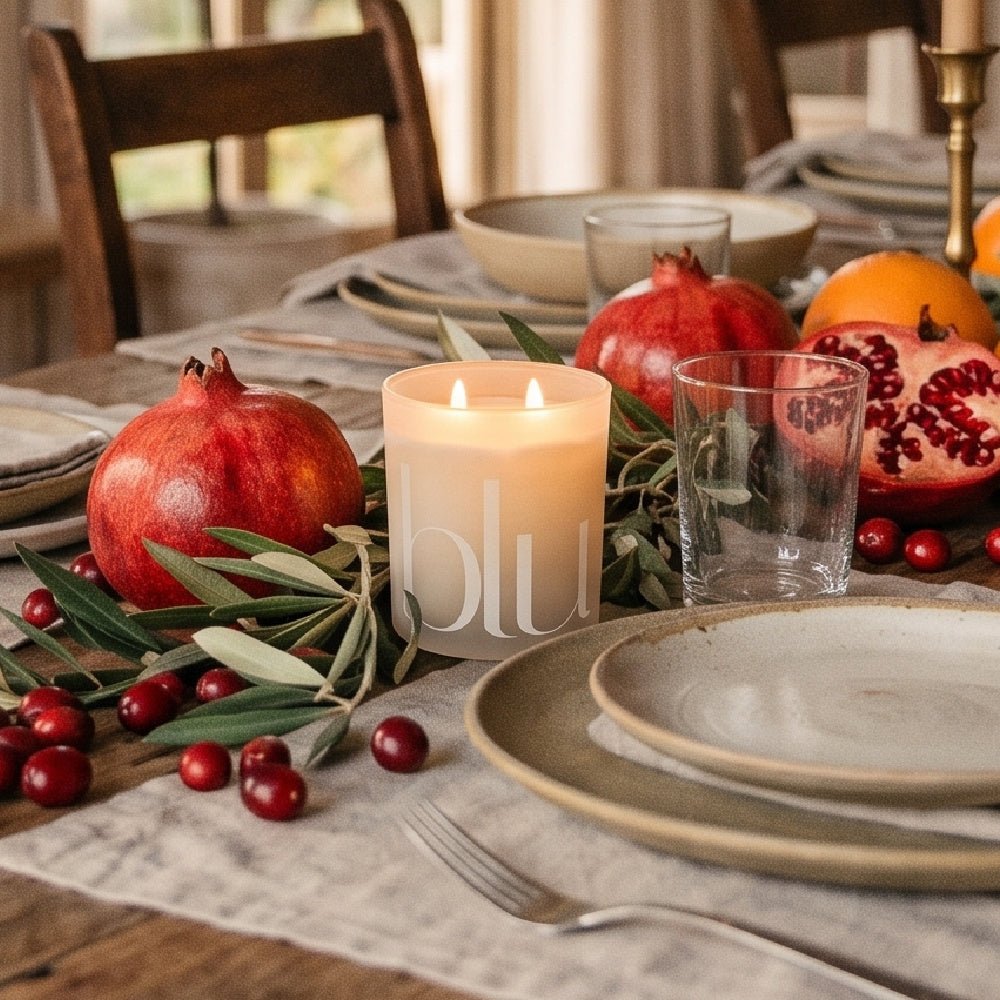 Gather artisan candle in frosted ivory glass with dual glowing flames centered on an autumn tablescape with pomegranates, fresh fruit, and olive branches