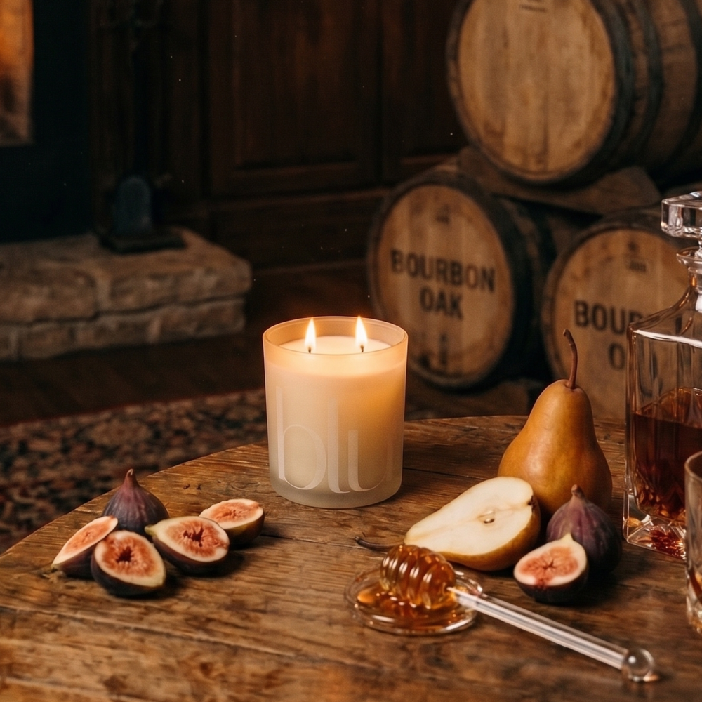 A lit white candle in a clear glass jar sits on a rustic wooden table, surrounded by sliced figs, a pear, a honey dipper, and a glass decanter, with stacked bourbon oak barrels in the background. figment artisan candle