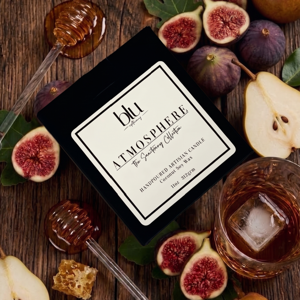 A square black candle tin labeled 'blu atmosphere The Sensuous Collection' is arranged on a rustic wooden surface with figs, honey, a glass of amber liquid with an ice cube, and honey dipper props. clean-burning fig bourbon coconut soy candle