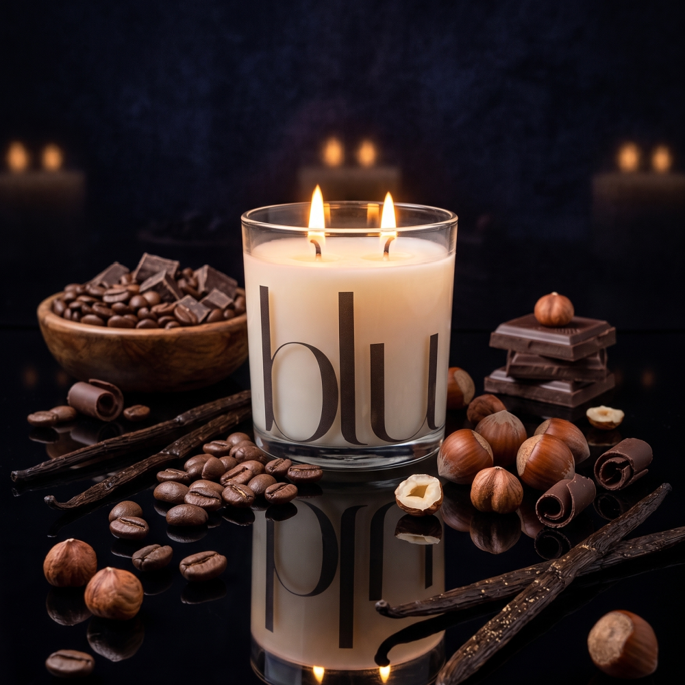 A double-wick candle in a clear glass jar with the word 'blu' printed on it, styled with coffee beans, chocolate pieces, vanilla sticks, and hazelnuts on a dark reflective surface against a dark background with soft blurred candles. clean-burning chocolate espresso coconut soy candle