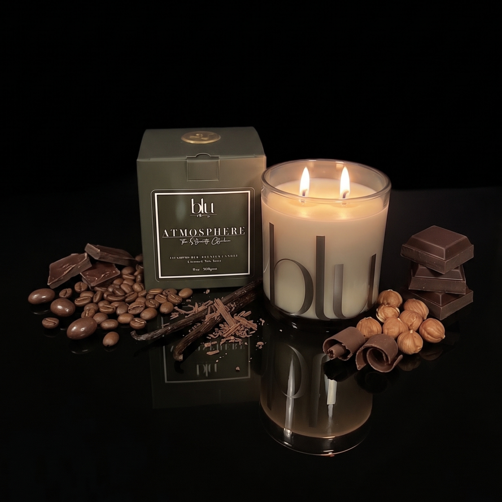 A lit double-wick candle in a clear glass jar sits beside its olive green box on a glossy black surface, surrounded by chocolate pieces, coffee beans, and cocoa nibs. The candle and box both show the brand BLU and the scent name ATMOSPHERE. hand-poured candle