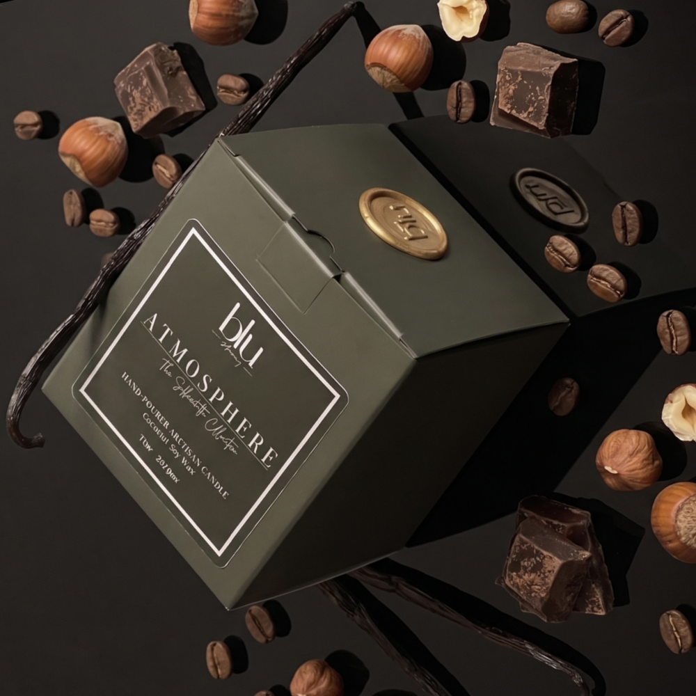 An olive green square gift box labeled 'blu Atmosphere' surrounded by chocolate pieces, coffee beans, and vanilla pods on a dark background. sultry gourmand candle