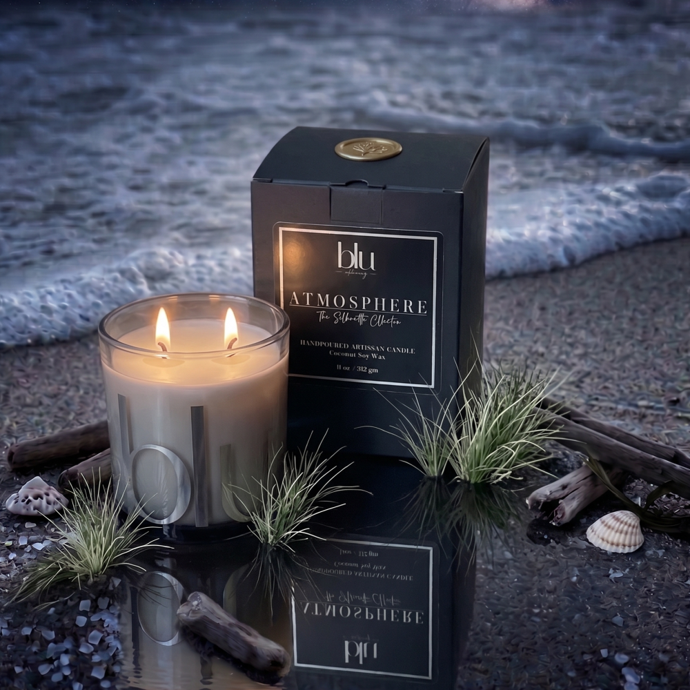 A lit atmospheric candle in a clear glass jar sits beside its black branded box on a reflective beach surface, with driftwood, seaweed, shells, and ocean waves in the background. Seaside Artisan Candle