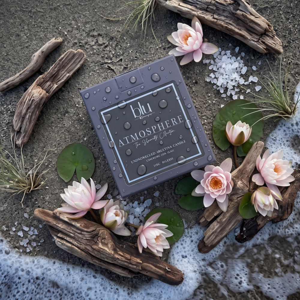 A square gray candle box labeled 'blu Atmosphere' is arranged on a beach-like surface with driftwood, water lilies, green lily pads, and white sea foam. The box is the main product, with decorative natural and aquatic props around it. sea salt coconut soy candle
