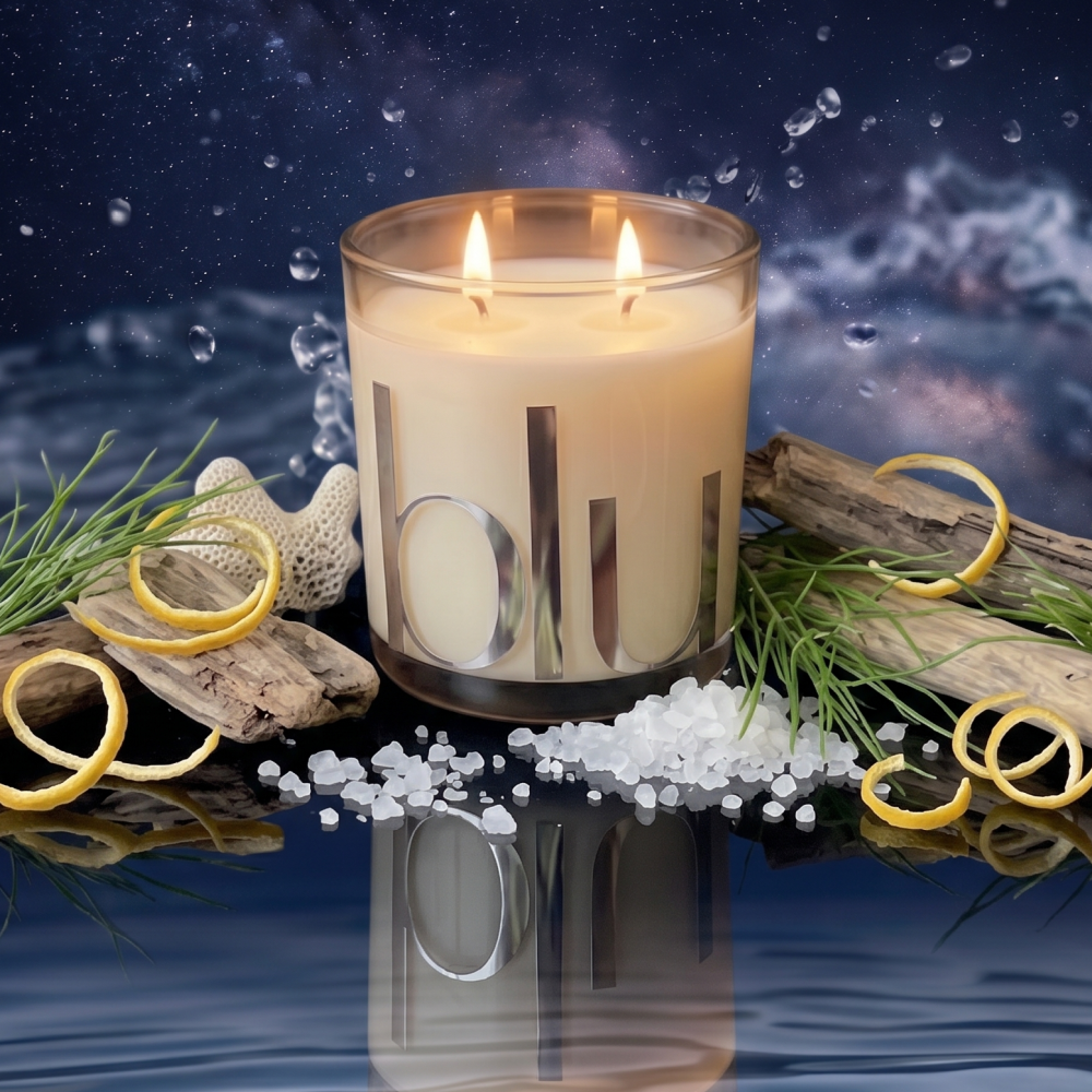 A double-wick candle in a clear glass jar with metallic vertical lettering, styled with sea salt, driftwood, greenery, and lemon curls against a dark water-like background with bubbles. sea salt coconut soy candle