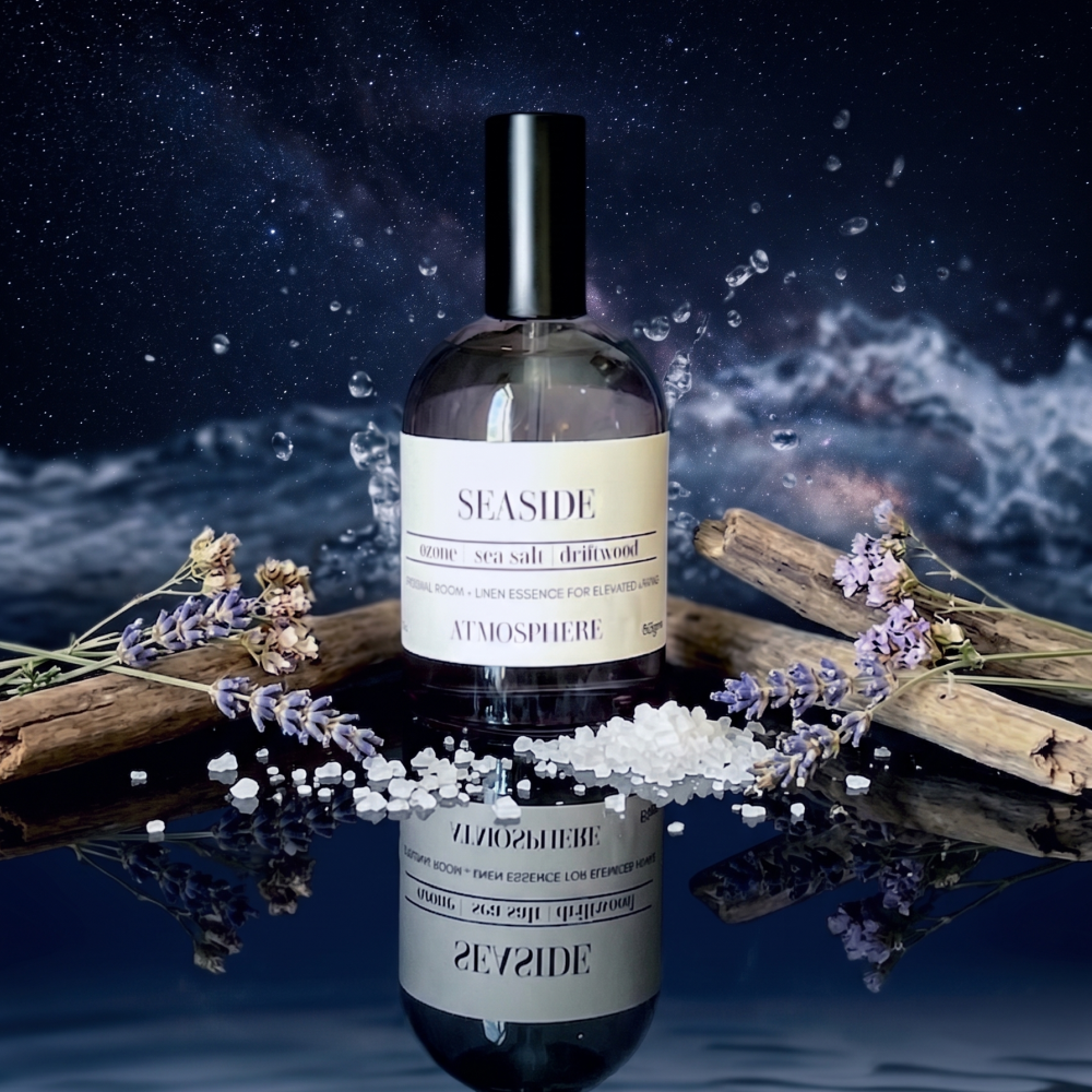 A bottle of Seaside sea salt drizzle room spray stands on a reflective dark surface with water droplets and dried lavender sprigs around it. The background shows a dramatic dark blue and black starry sky with splashing water, and the bottle is mirrored below. Seaside Room Spray
