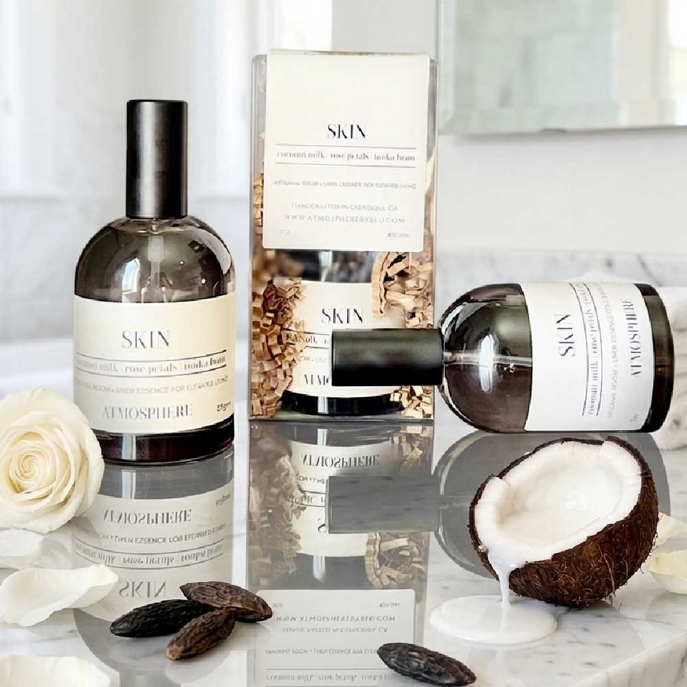 SKIN by Scent of the Sea Atmosphere body care set, featuring a dark glass bottle, a horizontal bottle, and a clear box with botanical imagery, styled on a marble surface with a coconut, rose petals, and dark chocolate. Skin Room Spray