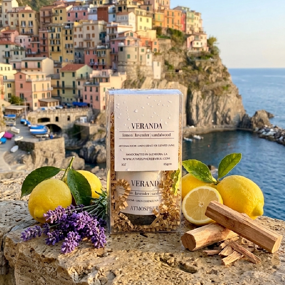 A clear rectangular box of Veranda lemon balm and lavender-infused steamers sits on a stone ledge outdoors, surrounded by lemons, lavender flowers, cinnamon sticks, and dried botanicals. The background shows a coastal cliffside town with blue sea and sky. Veranda Room Spray
