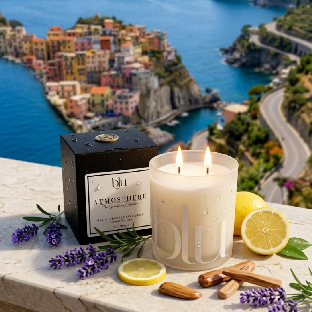 A lit double-wick scented candle in a frosted glass jar sits on a stone ledge with a black branded box beside it, surrounded by lavender sprigs, lemon halves, cinnamon sticks, and green leaves. The background shows a coastal town and blue sea, making this a lifestyle scene rather than a clean studio product shot. Veranda Artisan Candle
