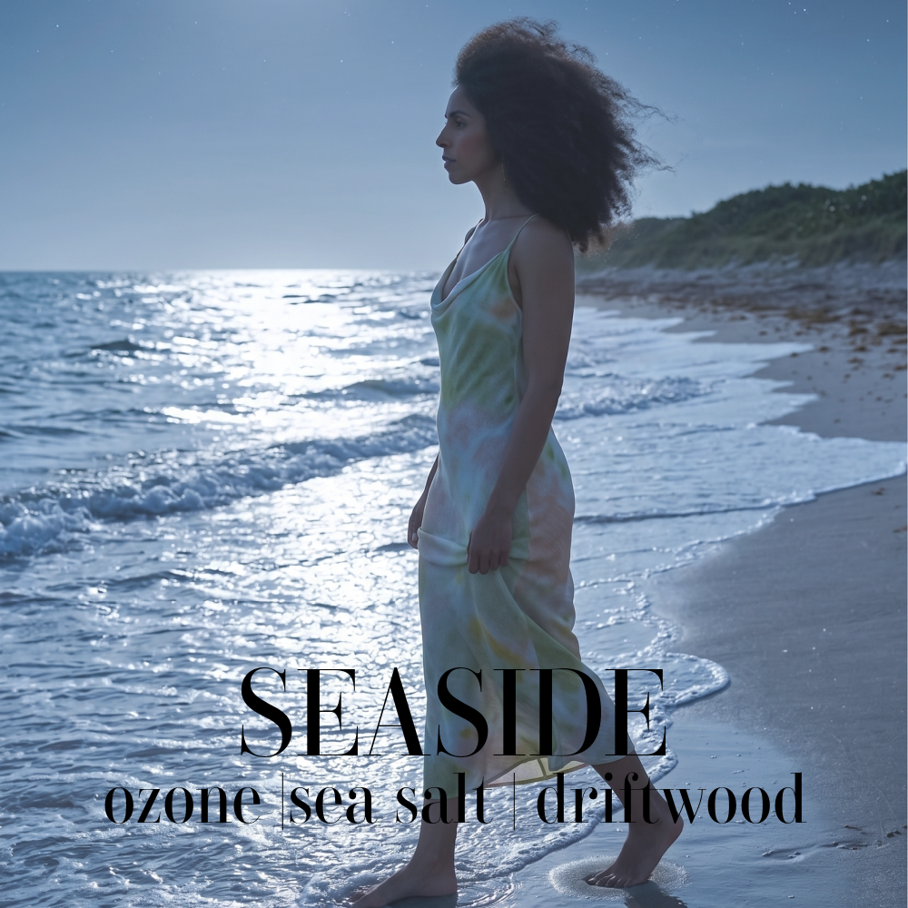 A woman stands in shallow ocean water on a beach at dusk, wearing a flowing pale green and cream satin dress. Large overlaid text reads 'SEASIDE' with smaller text 'ozone | sea salt | driftwood'. Seaside Artisan Candle